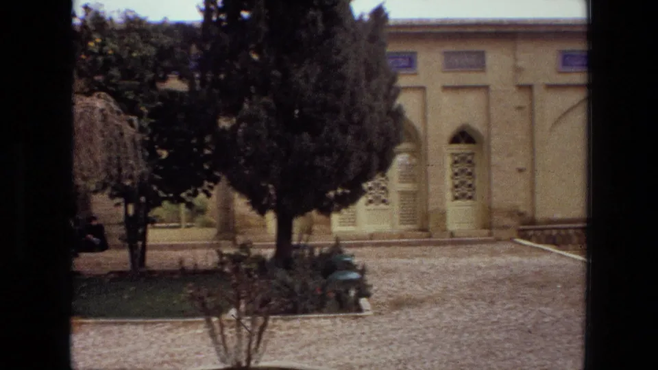 1972 Iran 8mm frame showing a pale facade with a doorway, mature trees, garden edging and a bench in the courtyard.