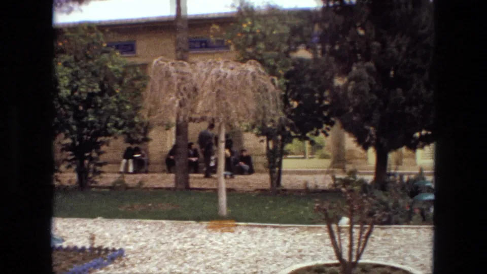 1972 Iran 8mm frame showing visitors gathered in shade near trees, lawn and a historic arched building facade.