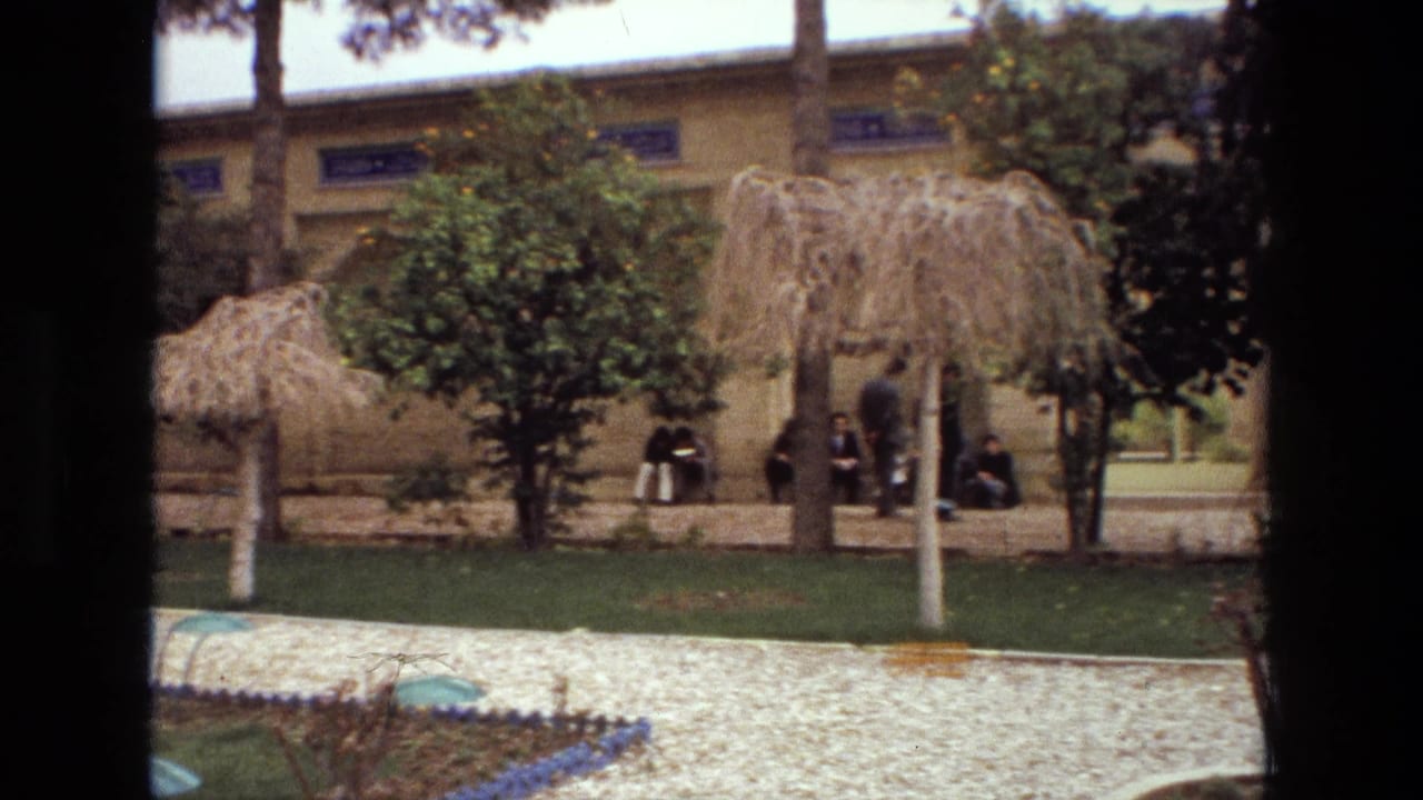 1972 Iran home-movie frame showing a walled garden with thatched shade structures, trees, lawn, gravel path, and visitors along the wall.