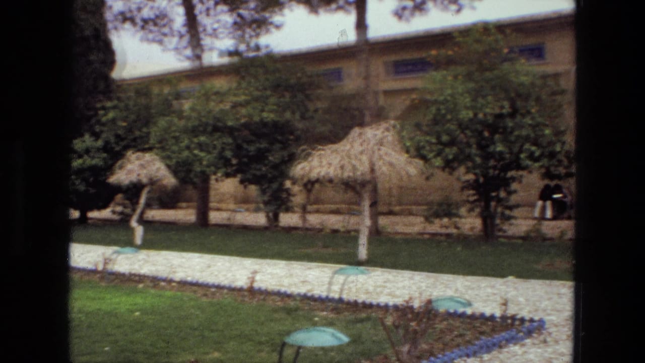 1972 Iran garden view with trees, thatched umbrellas, a tan perimeter wall, gravel path, lawn, and a green dome-shaped foreground fixture.