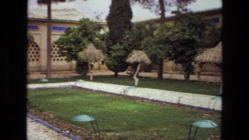 1972 Iran garden frame showing intersecting gravel paths, lawn, trees, thatched umbrellas, wall details, and green dome-shaped foreground fixtures.