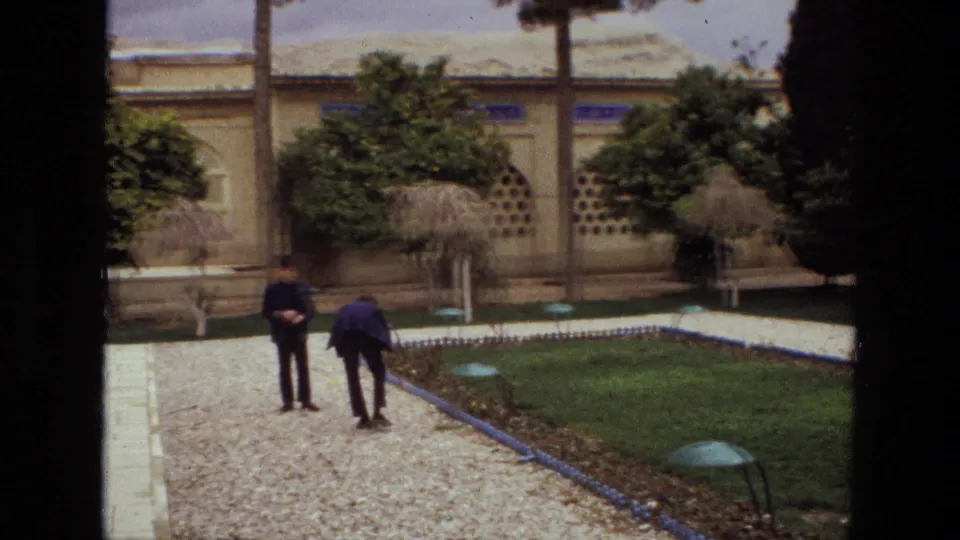 1972 Iran home-movie frame with two visitors walking along a gravel path in a walled garden with trees, lawn, and blue tile edging.