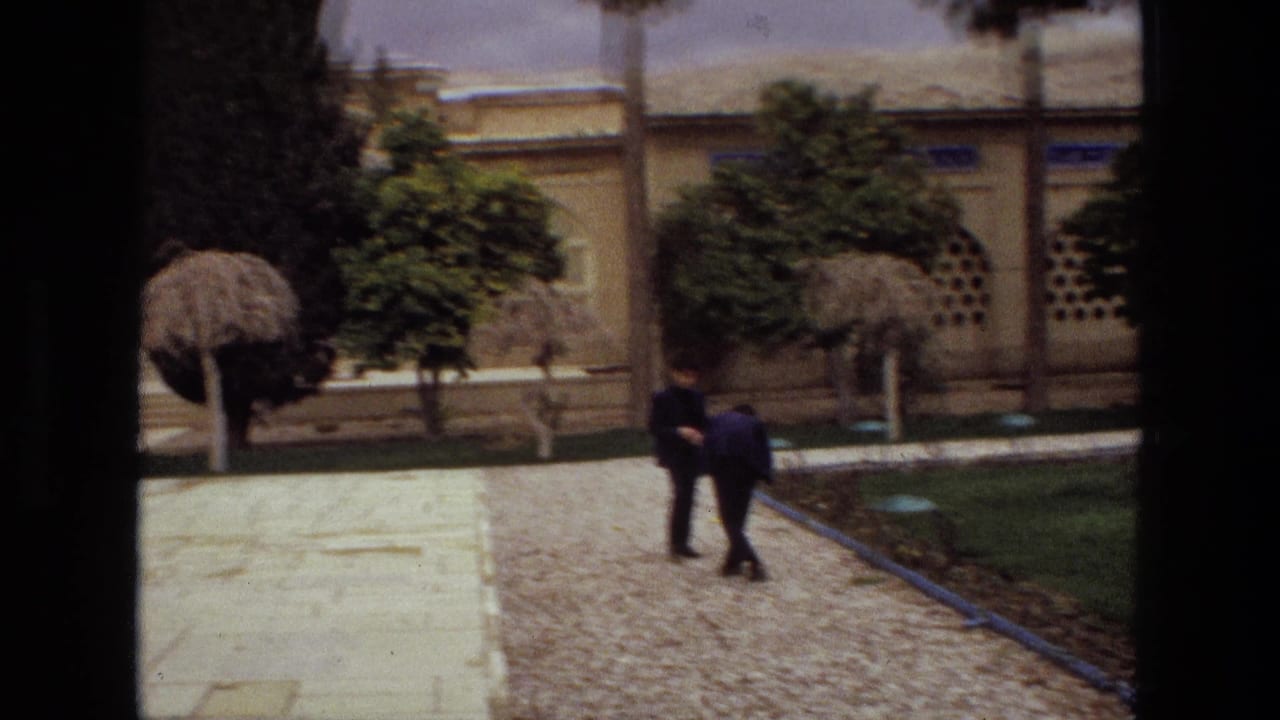 Courtyard with pale facade, trimmed trees, gravel path, lawn, and two people walking.