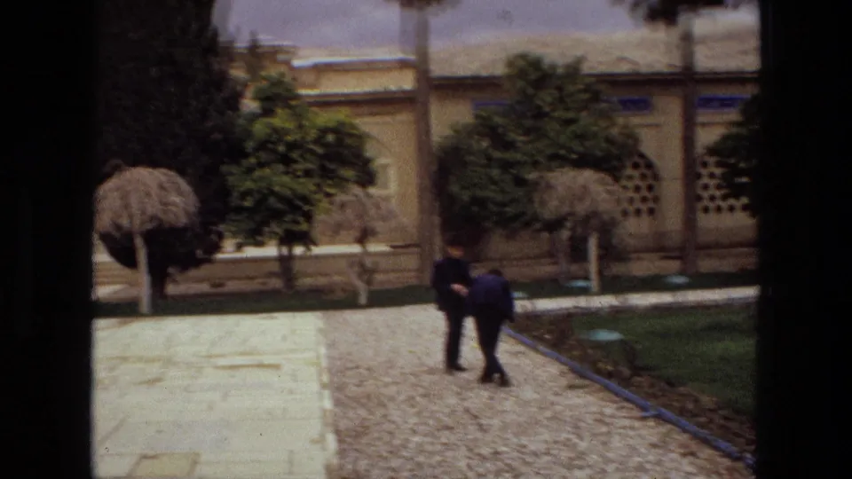 1972 Iran home-movie frame showing two visitors walking through a religious courtyard with manicured trees and a pale building facade.