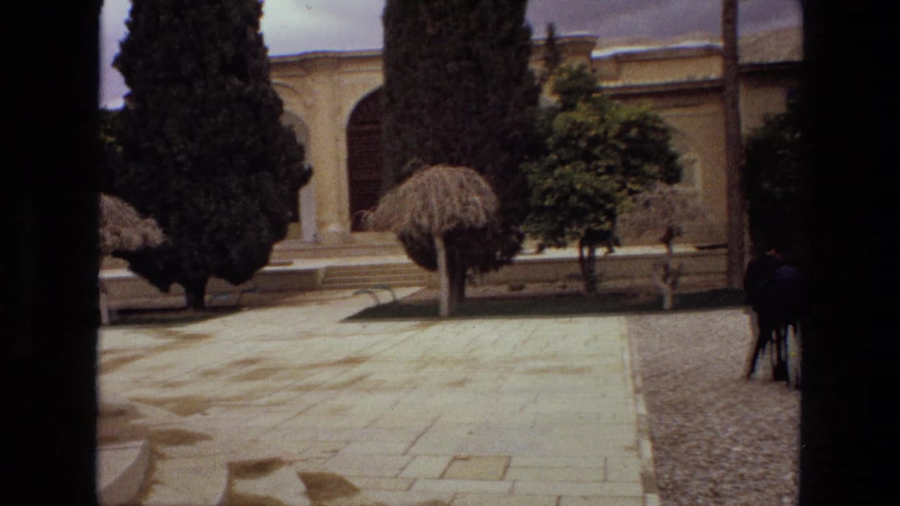 1972 Iran courtyard view with stone paving, gravel border, shaped trees, and distant visitors near a religious building facade.
