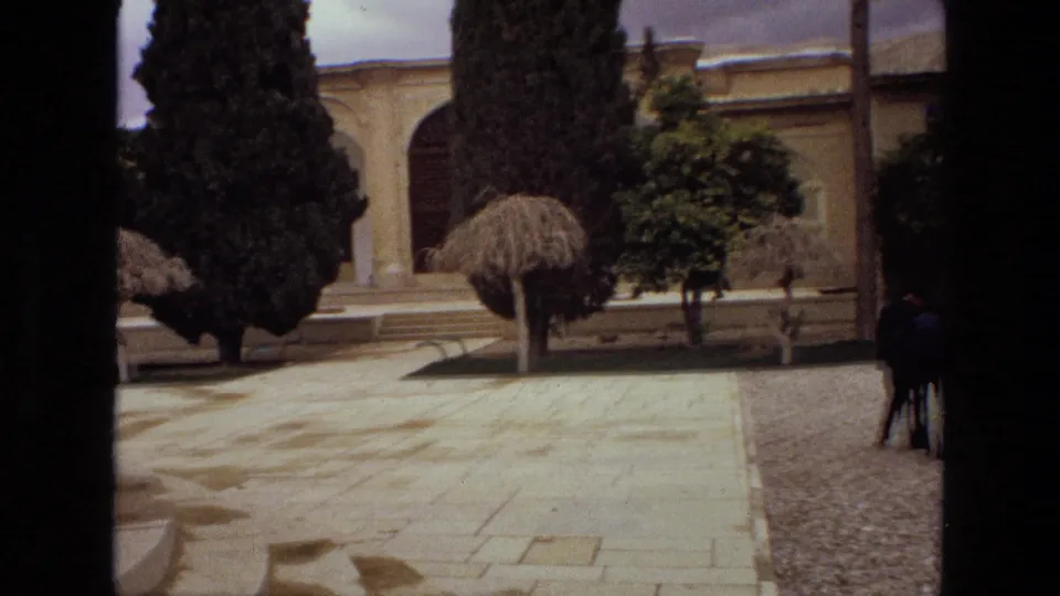 1972 Iran courtyard view with stone paving, gravel border, shaped trees, and distant visitors near a religious building facade.