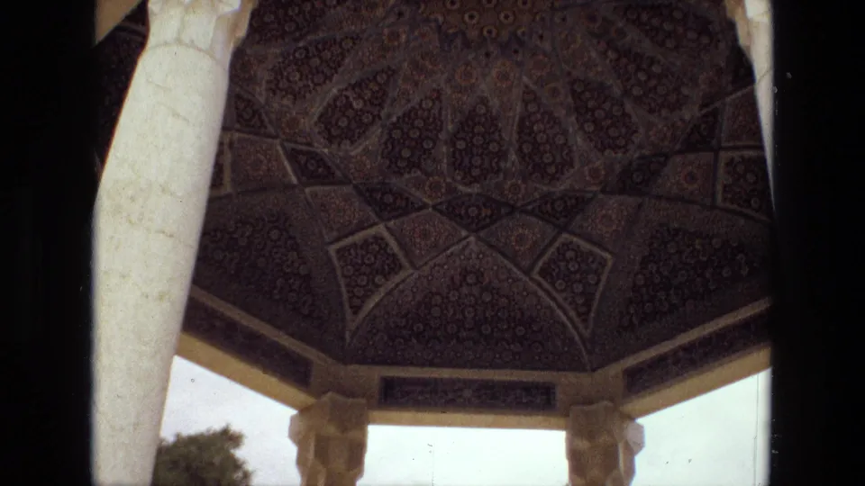 1972 Iran frame showing the underside of an ornate dome, pale supporting columns, carved capitals, and outdoor trees beyond.