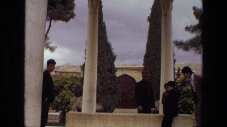 1972 Iran 8mm frame with men in dark clothing near a columned pavilion, garden wall, and tall cypress trees.
