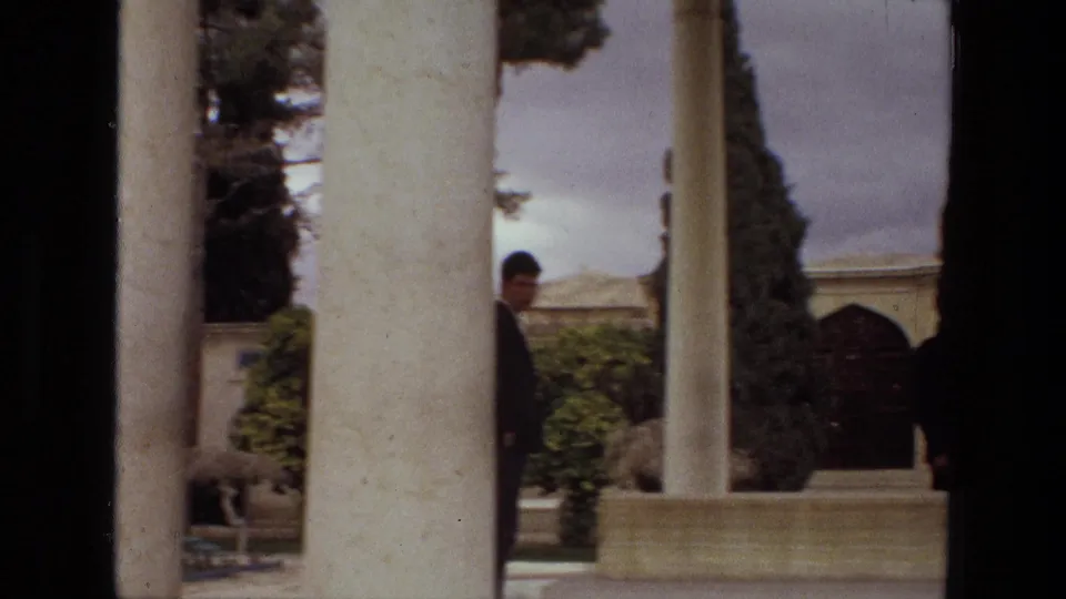 1972 Iran home-movie frame showing a man partly obscured by a column and another figure near a garden pavilion.