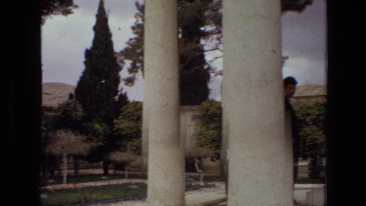 1972 Iran 8mm frame showing a courtyard seen through pale columns, with conifer trees, greenery, gravel paths, and hills beyond.