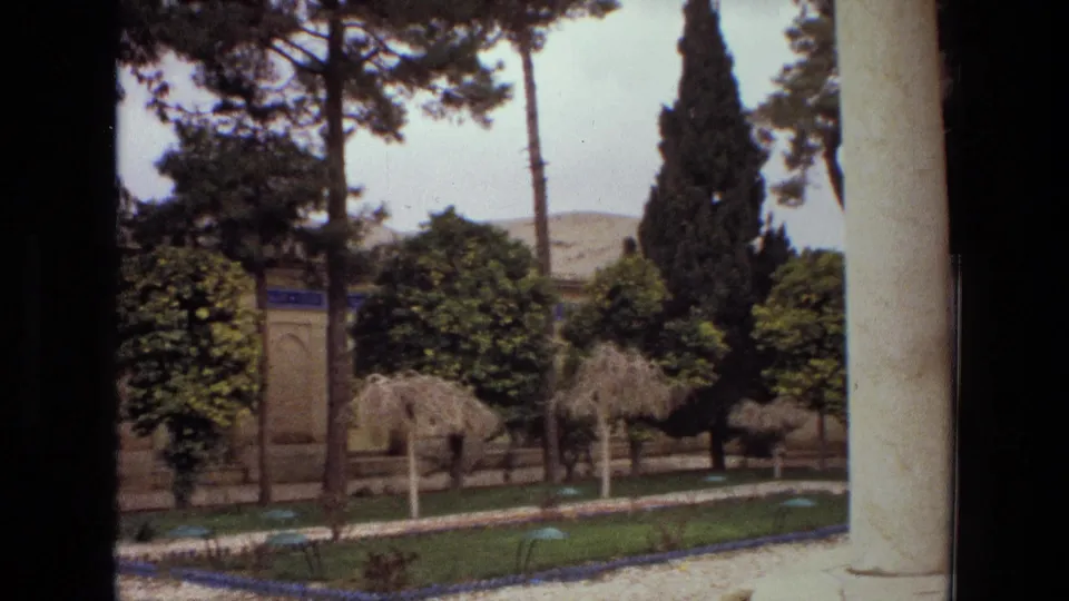 1972 Iran courtyard frame with tall trees, clipped shrubs, a gravel path, low garden borders, and a blue tile panel on the building wall.