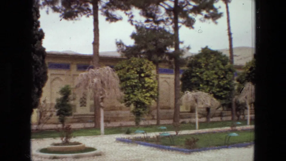 1972 Iran 8mm frame showing gravel paths, shaped garden trees, green lawn beds, tall trunks, and traditional architectural wall details.
