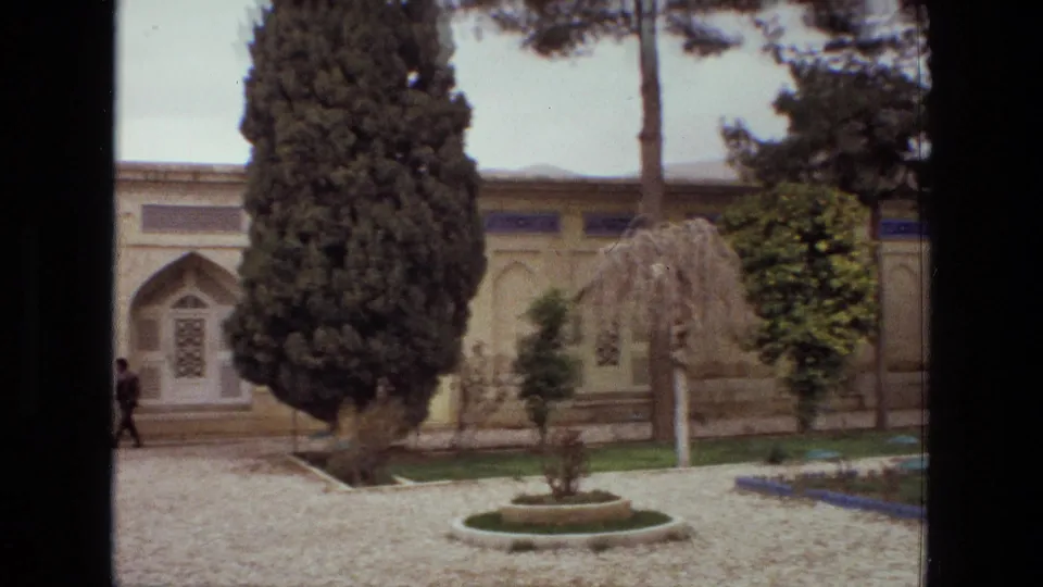 1972 Iran frame with a gravel path, arched doorway, tall conifer, circular planter, lawn borders, and a distant walking figure.