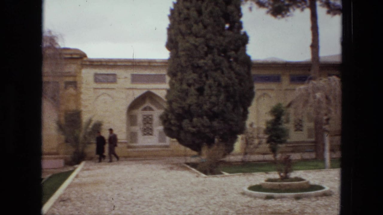 Two people walk across a courtyard in front of an arched building entrance and tall trees.