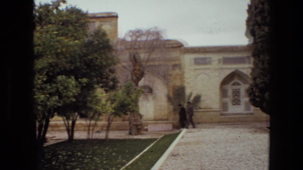 1972 Iran frame of a landscaped courtyard with two distant visitors near arched architecture, trimmed lawn borders, and leafy trees.
