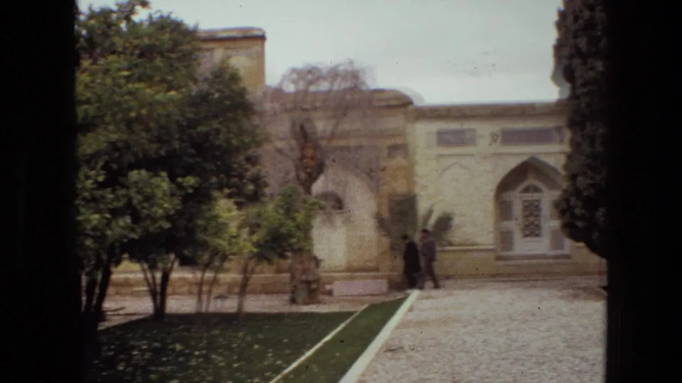 1972 Iran frame of a landscaped courtyard with two distant visitors near arched architecture, trimmed lawn borders, and leafy trees.