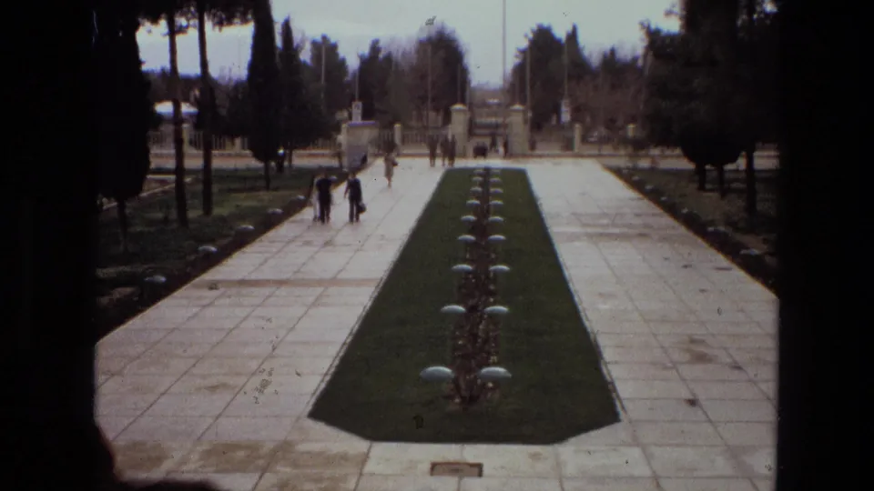 1972 Iran home-movie frame of a formal garden axis with white tiled paths, a central grass strip, low plantings, trees, and distant visitors.
