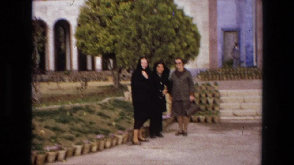 The three women appear farther from the camera in an Iranian garden, with steps, planted pots, trees, and a building entrance visible.