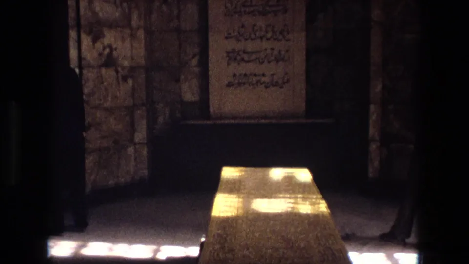 1972 Iran footage frame showing a stone tomb beneath a wall panel with Arabic-script writing in a dim interior.