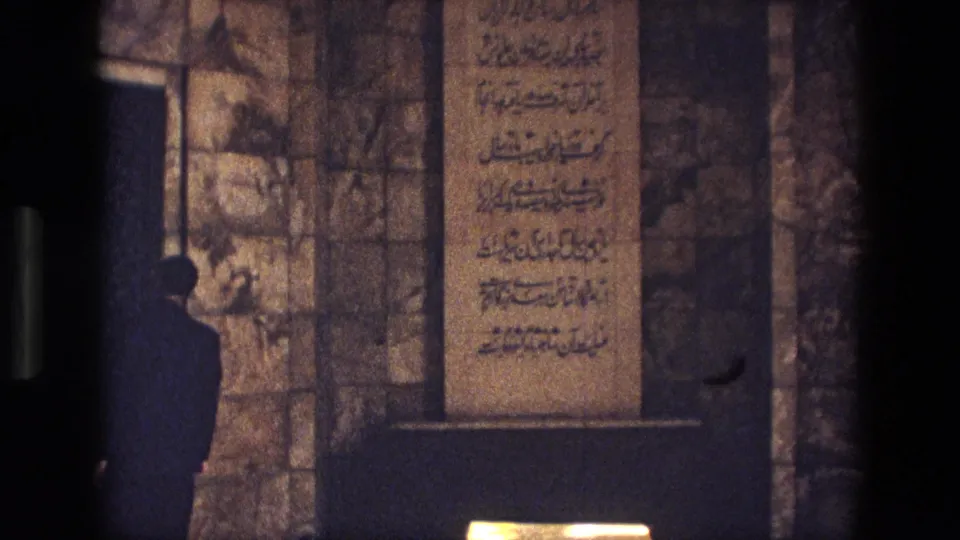A shadowed visitor stands near a stone tomb and vertical Arabic-script inscription panel in a historic Iranian interior.