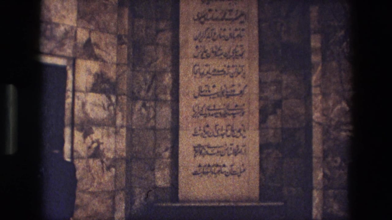1972 Iran 8mm frame showing a calligraphic stone tablet on an interior wall, with a dark-clothed adult partly visible at left.