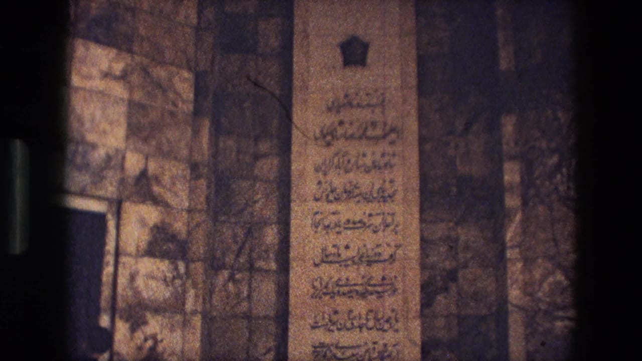 1972 Iran 8mm frame of an inscribed stone panel in a dim historic interior with surrounding masonry and an arched recess.