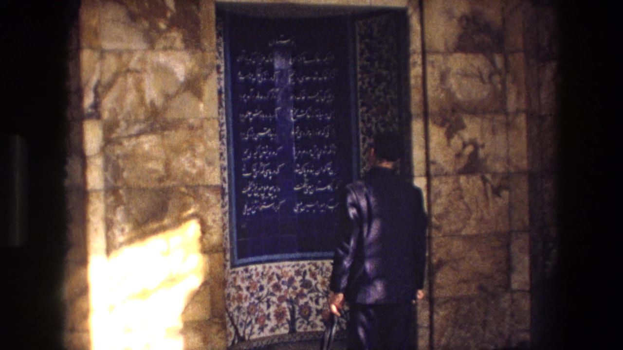 Man in a dark suit stands before a blue Persian calligraphy panel.