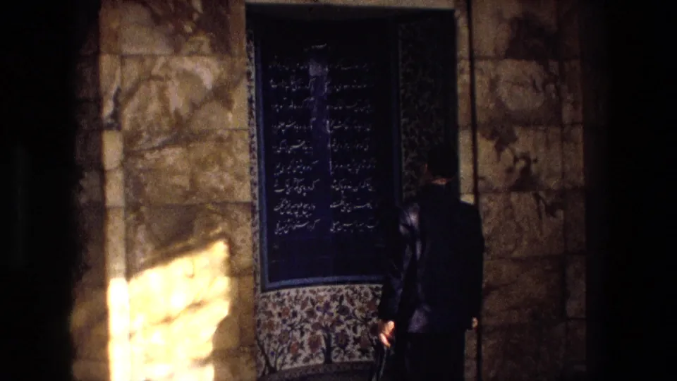 From a handheld angle, a man stands near a blue Persian calligraphy panel and patterned tilework in Iran.