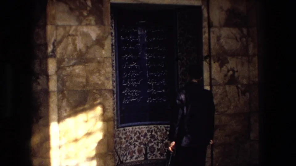 A suited man stands partly turned beside a blue Persian writing panel and stone wall in 1972 Iran 8mm footage.