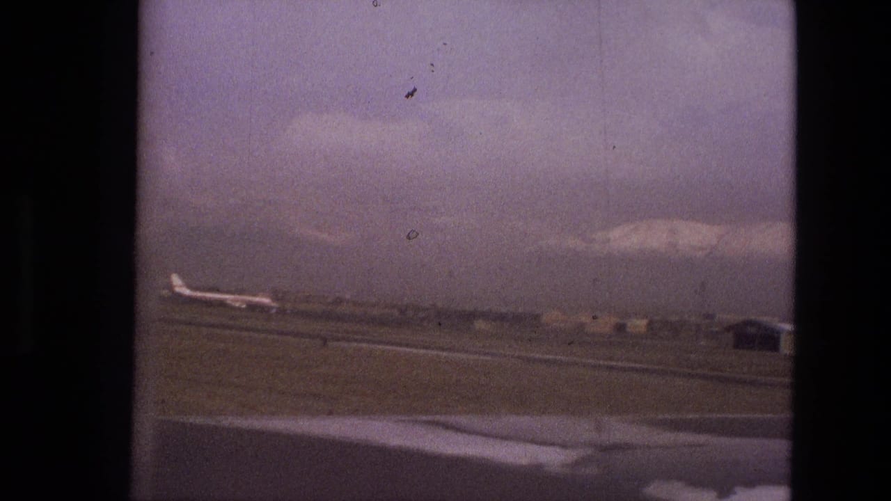 Airport tarmac seen through a window with a distant jet and mountains beyond.