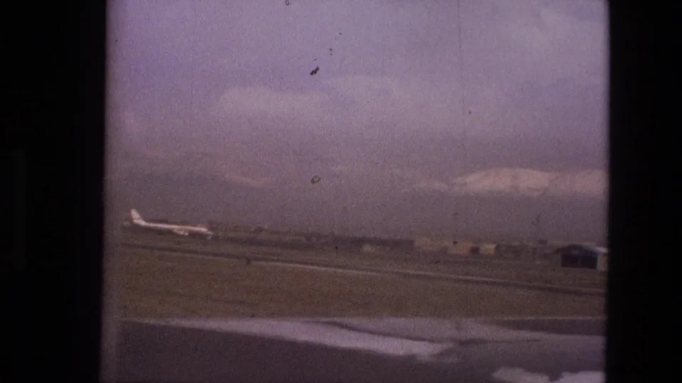 1972 Iran airport view from indoors, showing a tarmac, distant airplane, low buildings, and mountains under a hazy sky.