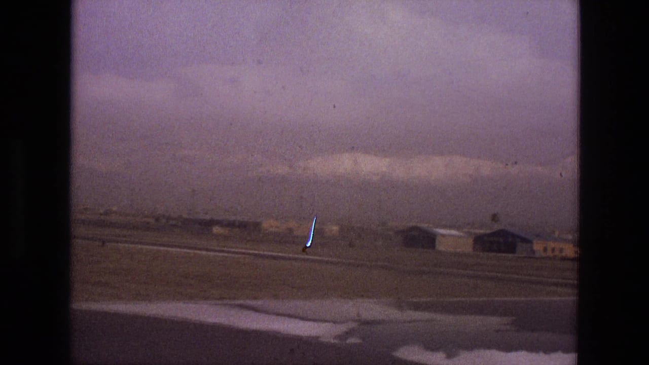 View across an Iranian airport in 1972, with runway or taxiway lines, hangars, and snow-capped mountains beyond the field.