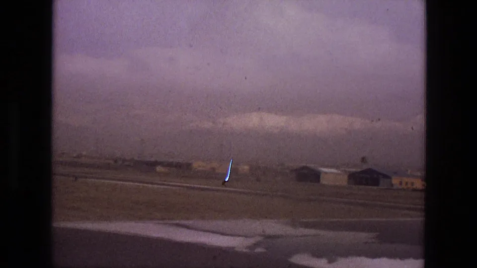 View across an Iranian airport in 1972, with runway or taxiway lines, hangars, and snow-capped mountains beyond the field.