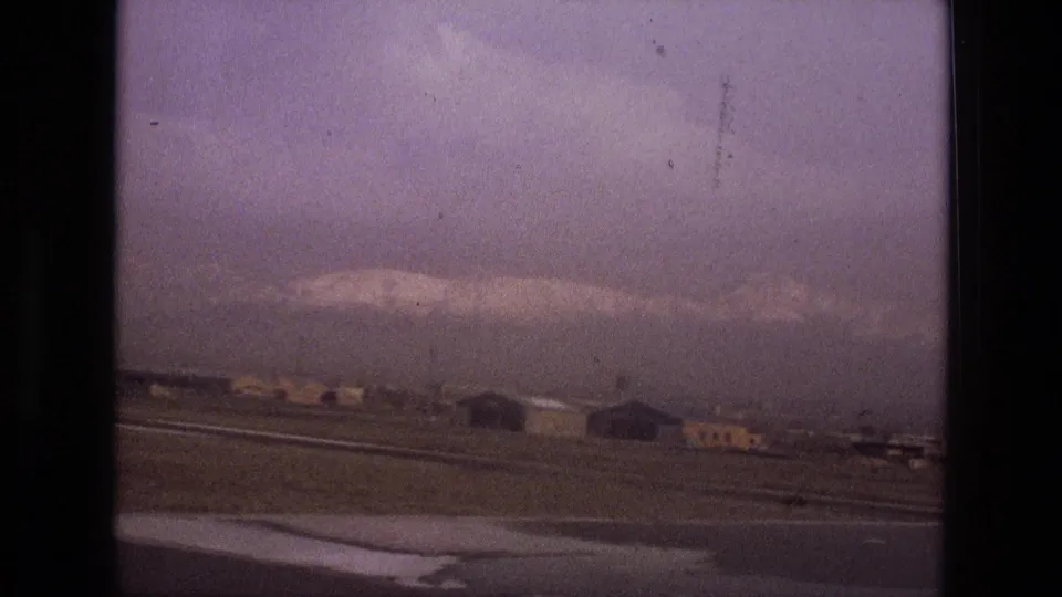 1972 Iran airport scene with low buildings near the runway and a hazy mountain range visible beyond the airfield.