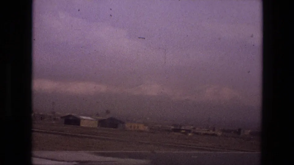 Terminal-window view at an Iranian airport in 1972, with an airplane wing edge, tarmac, hangars, and distant mountains.