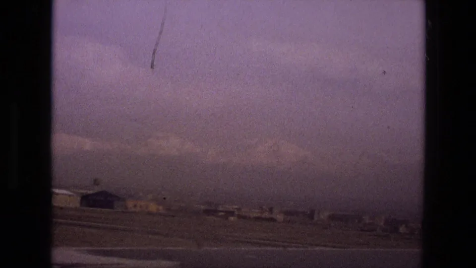 1972 Iran 8mm frame showing a hazy urban edge with low buildings, open foreground, distant terrain, and dark film gate borders.