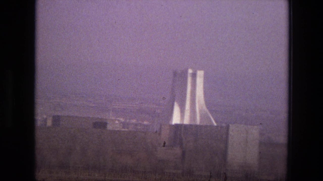 1972 Iran 8mm frame with a distant modernist twin-tower landmark rising behind low buildings under a hazy purple sky.