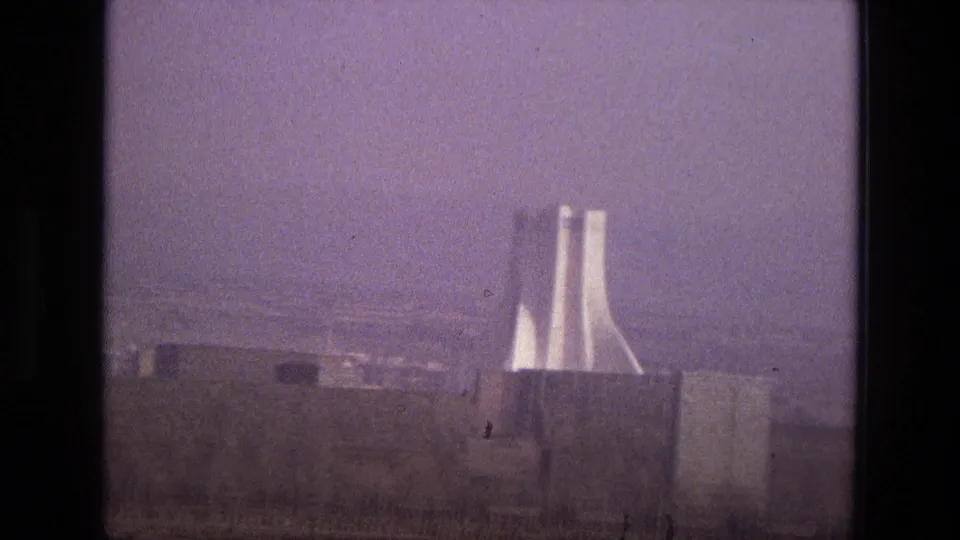 1972 Iran 8mm frame with a distant modernist twin-tower landmark rising behind low buildings under a hazy purple sky.