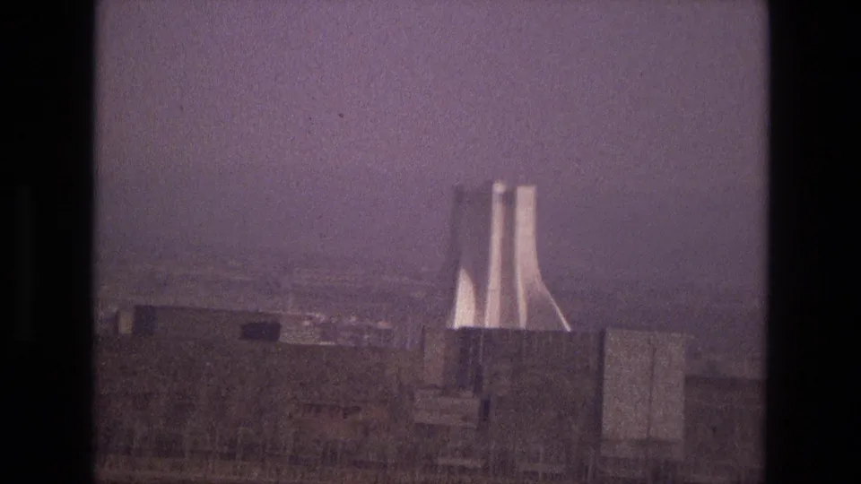 1972 Iran 8mm frame centering a modernist twin-tower landmark beyond rooftops, scrubby foreground, and a hazy city backdrop.