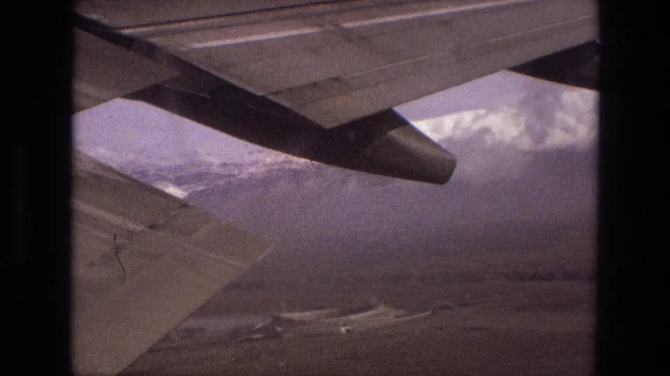 1972 Iran 8mm passenger-window view with an aircraft wing above hazy snow-capped mountains and rugged terrain below.