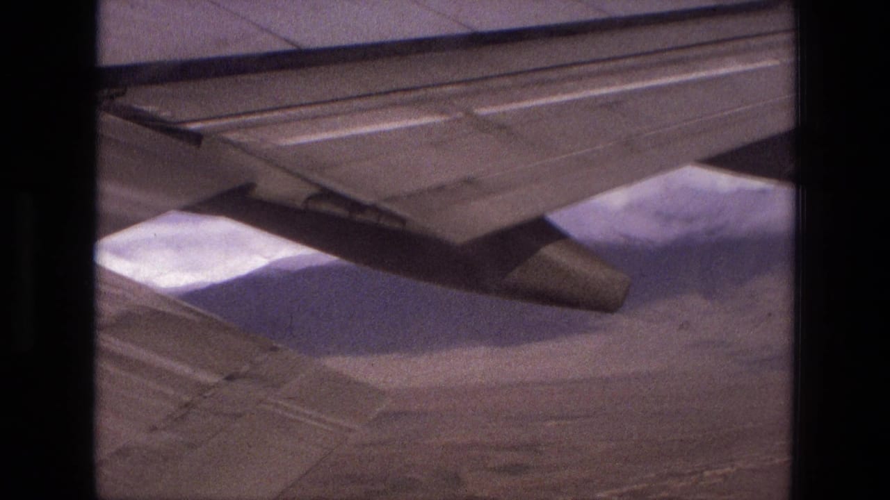 Aircraft wing and engine over pale mountains and arid ground.
