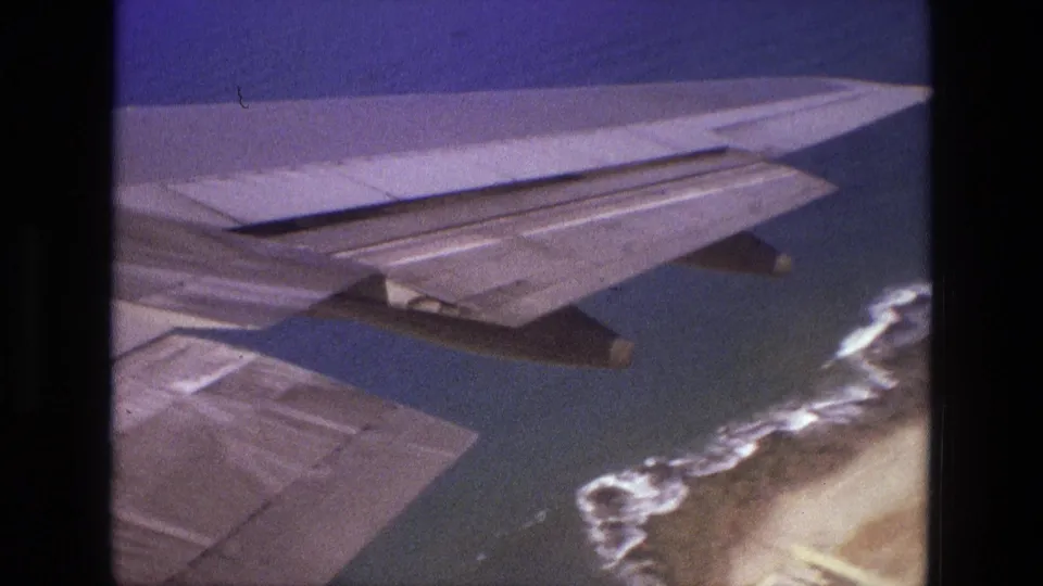 1972 Iran aerial home-movie frame showing an airplane wing over blue-green water, surf, and a sandy coastal edge.