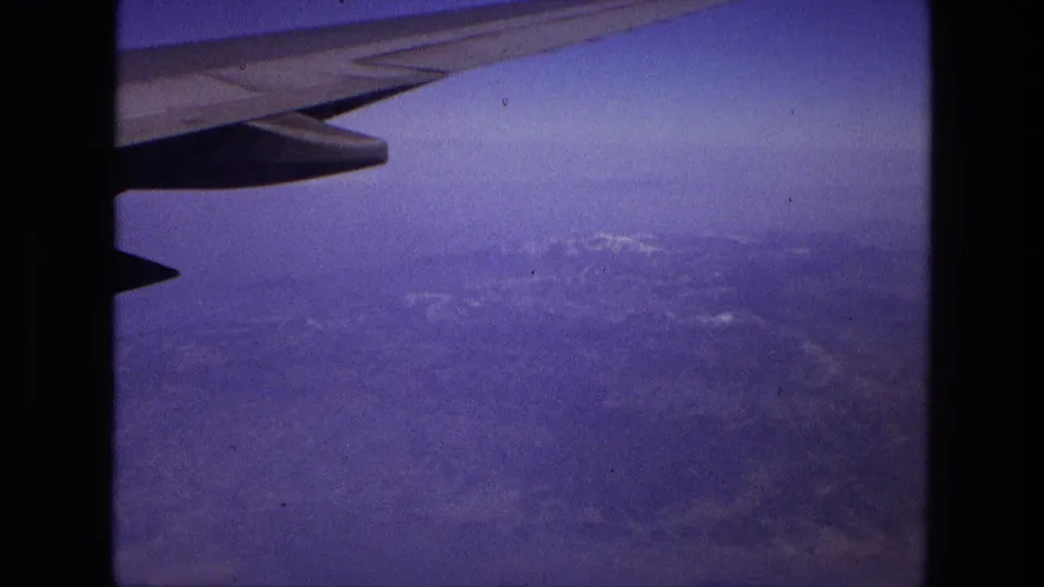 1972 8mm frame from an airplane window in Iran, with the wing at top and distant rugged land below a hazy sky.