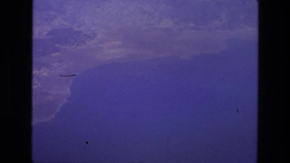 1972 8mm frame from Iran showing a coastline and open water through an airplane window, with haze and film specks visible across the scan.