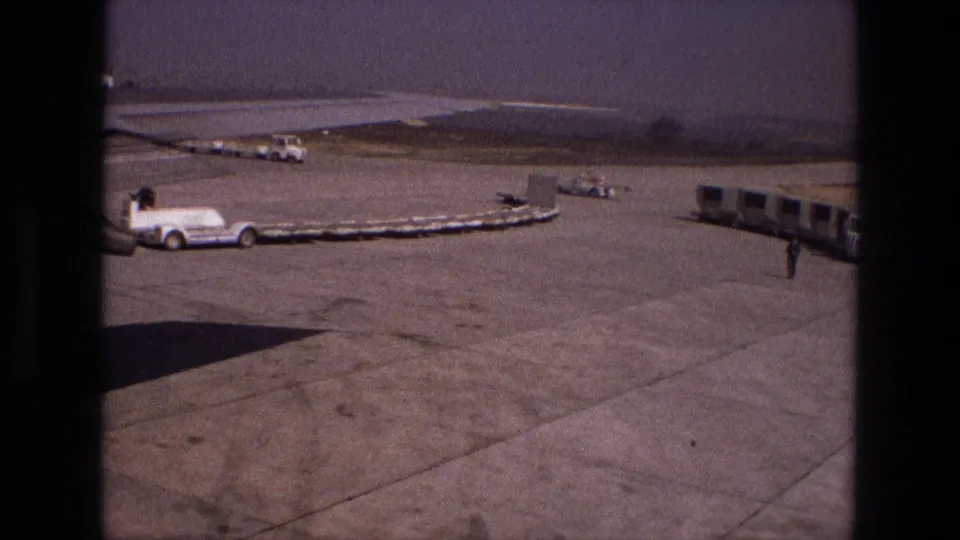 Iran 1972 tarmac view with a long cargo belt, white service vehicle, mobile passenger stairs and a distant worker near ground equipment.