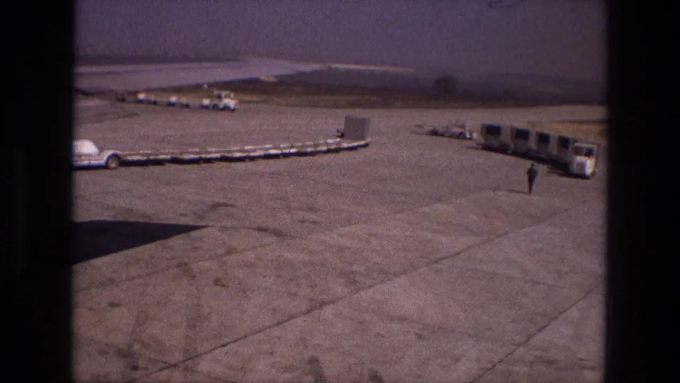 Iran 1972 airport apron from a passenger viewpoint, showing cargo handling equipment, service vehicles, mobile stairs and a distant ground worker.