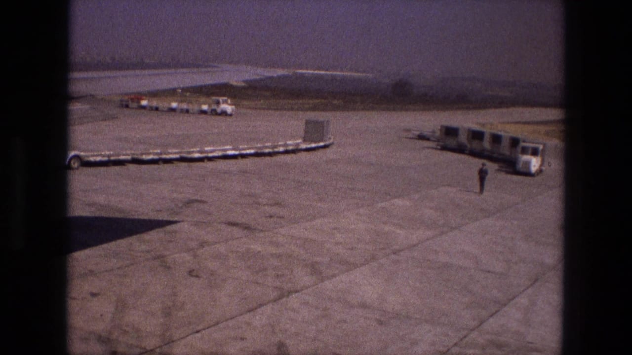 Airport tarmac with linked luggage carts, distant vehicles, and a lone worker near a passenger bus.