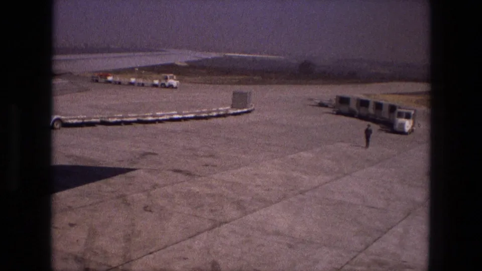 1972 Iran airport tarmac seen from behind a dark window edge, with luggage carts, distant vehicles, and a ground worker near a mobile passenger bus.