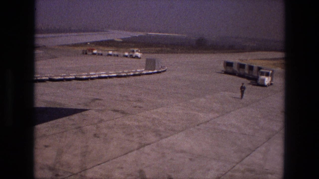 1972 Iran airport ground scene with a long cart line, a mobile passenger bus, distant service vehicles, and one worker crossing the open tarmac.