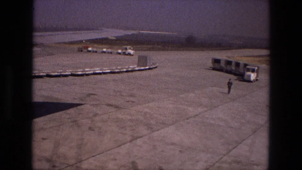 1972 Iran airport ground scene with a long cart line, a mobile passenger bus, distant service vehicles, and one worker crossing the open tarmac.